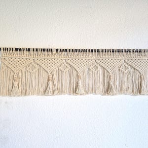 May include: A cream-colored macrame wall hanging featuring a series of repeating triangular knot patterns. The piece is suspended from a thin black rod and includes vertical fringe sections accented with small wooden beads and tassels.