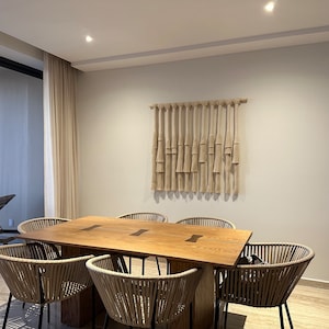 K&ouml;nnte beinhalten: Ein Esstisch aus Holz mit sechs geflochtenen St&uuml;hlen. Der Tisch hat eine nat&uuml;rliche Holzoptik und eine hellbraune Farbe. Die St&uuml;hle sind aus geflochtenem Rattan und haben schwarze Metallbeine. Eine gewebte Wandbehang h&auml;ngt an der Wand hinter dem Tisch.