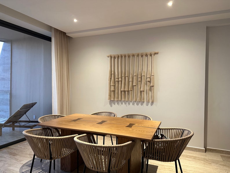 K&ouml;nnte beinhalten: Ein Esstisch aus Holz mit sechs geflochtenen St&uuml;hlen. Der Tisch hat eine nat&uuml;rliche Holzoptik und eine hellbraune Farbe. Die St&uuml;hle sind aus geflochtenem Rattan und haben schwarze Metallbeine. Eine gewebte Wandbehang h&auml;ngt an der Wand hinter dem Tisch.