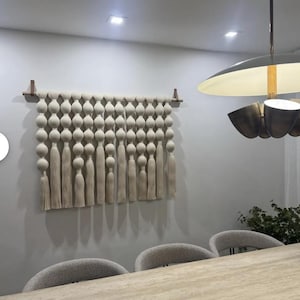 Könnte beinhalten: Ein weißer Makramee-Wandbehang mit einer Reihe von geknüpften Kugeln und Quasten. Der Behang ist an einer Wand mit einer Holzleiste befestigt.