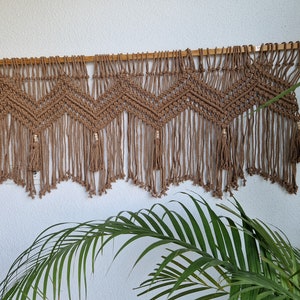 May include: A brown macrame wall hanging with a chevron pattern and tassels. The hanging is made of natural fiber rope and is attached to a wooden dowel.