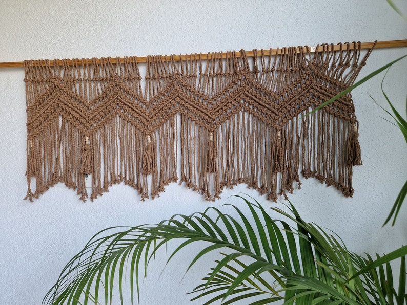 May include: A brown macrame wall hanging with a chevron pattern and tassels. The hanging is made of natural fiber rope and is attached to a wooden dowel.
