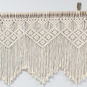 May include: An off-white macrame wall hanging featuring a series of repeating diamond-shaped knots and long, flowing fringe. The textile is suspended from a smooth, light-colored wooden rod, which is held in place by a small, decorative metal hook.