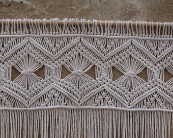 May include: White macrame wall hanging with a geometric pattern and fringe. The hanging is made of cotton cord and has a natural, bohemian style.