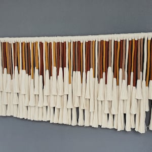 May include: A wall hanging made of white cotton yarn with brown, orange, and yellow accents. The hanging features a series of tassels that are arranged in a cascading pattern.