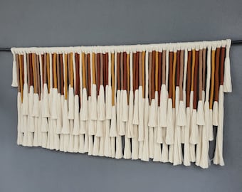 May include: A wall hanging made of white cotton yarn with brown, orange, and yellow accents. The hanging features a series of tassels that are arranged in a cascading pattern.