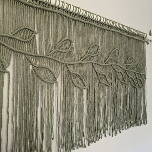 May include: A macrame wall hanging with a leaf design in a sage green color. The hanging is made with cotton cord and has a fringe at the bottom.