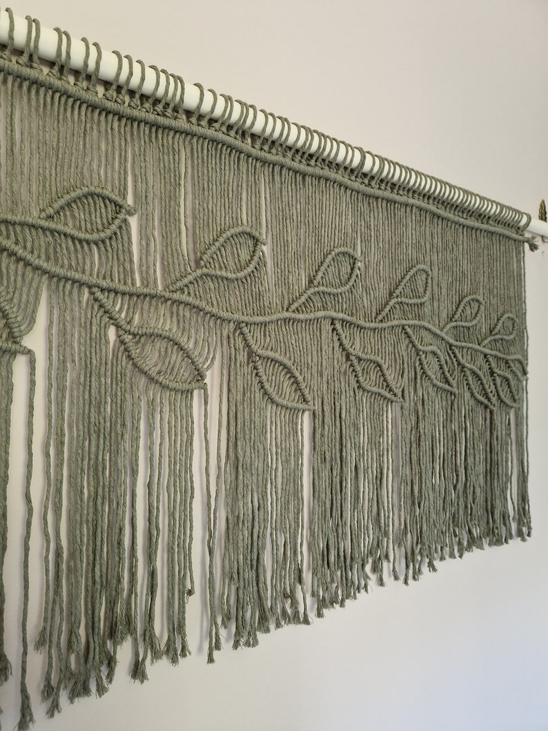 May include: A macrame wall hanging with a leaf design in a sage green color. The hanging is made with cotton cord and has a fringe at the bottom.
