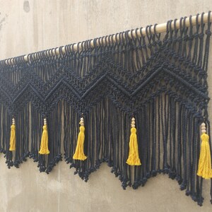 May include: Black macrame wall hanging with a geometric pattern and yellow tassels. The hanging is attached to a wooden dowel.