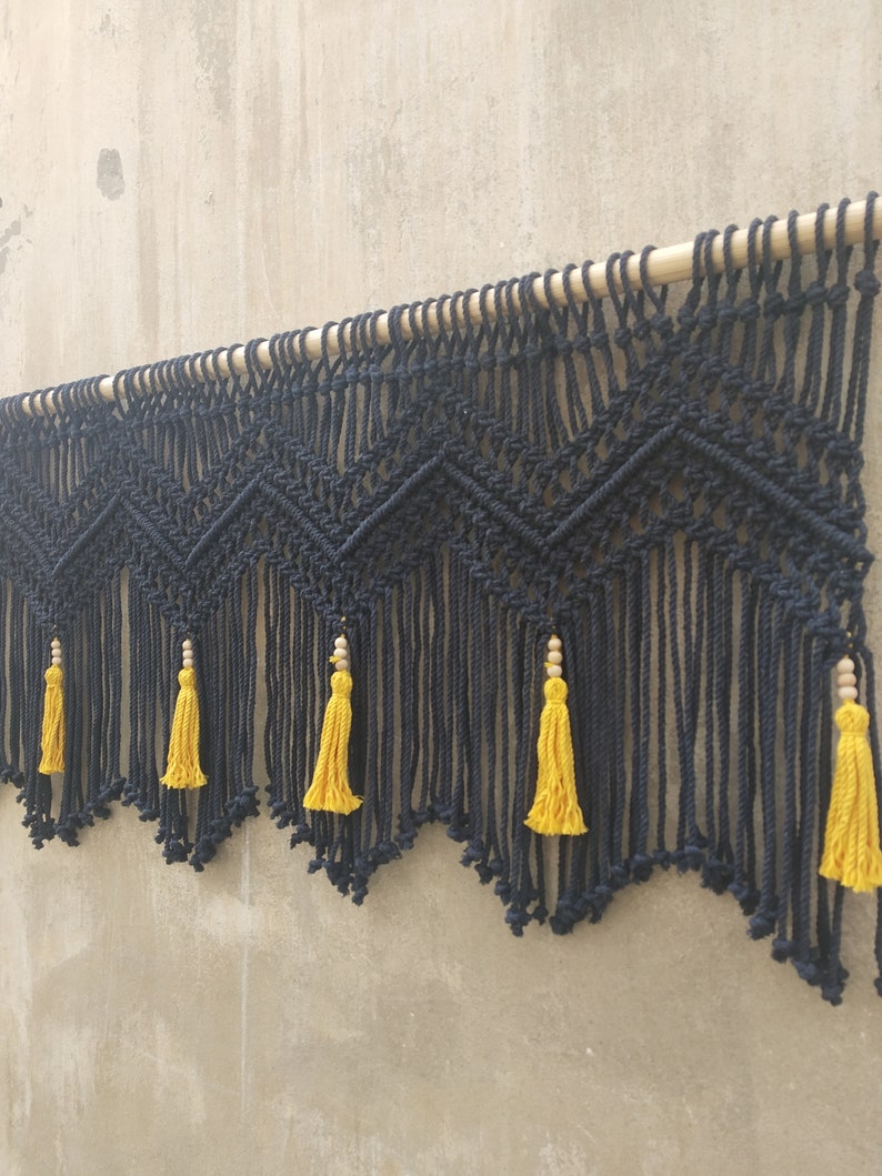 May include: Black macrame wall hanging with a geometric pattern and yellow tassels. The hanging is attached to a wooden dowel.