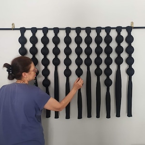 May include: Black wall hanging with a series of connected, rounded shapes and elongated tassels. The art piece is mounted on a black rod with gold-colored accents. The wall is painted white.