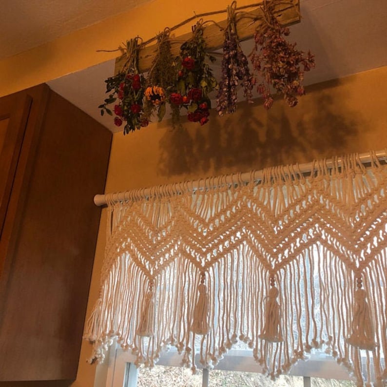 K&ouml;nnte beinhalten: Ein wei&szlig;er Makramee-Vorhang mit Chevron-Muster und Quasten h&auml;ngt &uuml;ber einem Fenster. Getrocknete Blumen h&auml;ngen an der Decke &uuml;ber dem Vorhang.
