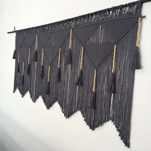 May include: Gray macrame wall hanging with wooden beads and tassels. The hanging is made of thick cotton cord and has a geometric pattern.