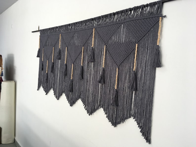 May include: Gray macrame wall hanging with wooden beads and tassels. The hanging is made of thick cotton cord and has a geometric pattern.