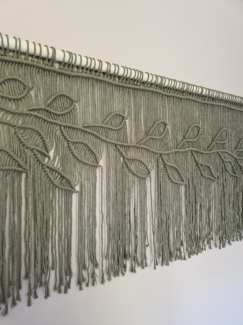 May include: A macrame wall hanging with a leaf design in a sage green color. The hanging is made of cotton cord and has a fringe at the bottom.