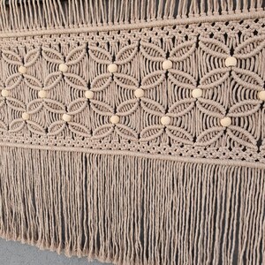 May include: Beige macrame wall hanging with a geometric floral pattern and wooden beads. The handcrafted piece features a fringe at the bottom and is mounted on a black rod. This decorative textile adds a bohemian touch to any space.