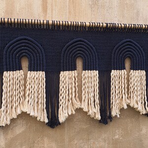 May include: A wall hanging with three black and white macrame arches. The arches are made with thick rope and have a fringe of the same rope at the bottom.