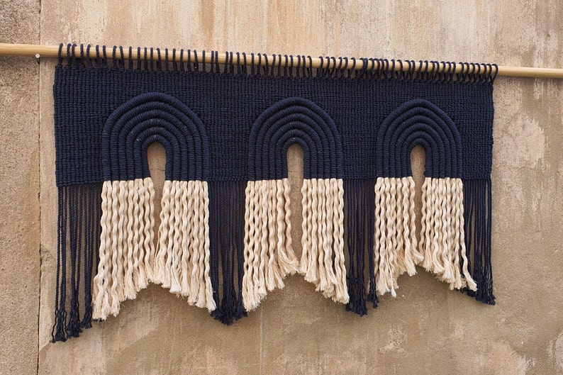 May include: A wall hanging with three black and white macrame arches. The arches are made with thick rope and have a fringe of the same rope at the bottom.