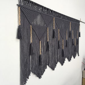 May include: A gray macrame wall hanging with wooden beads and tassels. The hanging is in a geometric pattern with a triangular shape.