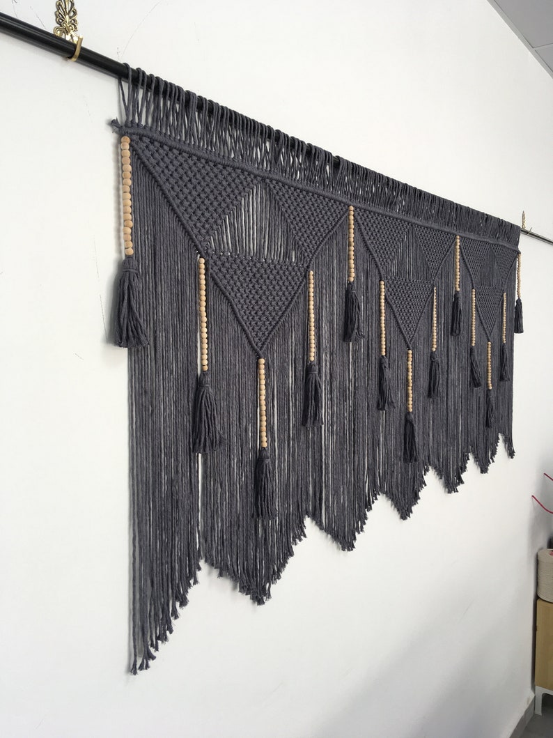 May include: A gray macrame wall hanging with wooden beads and tassels. The hanging is in a geometric pattern with a triangular shape.
