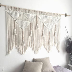 May include: A large off-white macrame wall hanging suspended from a wooden rod. The piece features intricate knotted diamond patterns, vertical fringe, and several wooden beads integrated into the design with small tassels at the ends of cords.