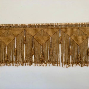 May include: A mustard yellow macrame wall hanging with a geometric pattern and white beads. The hanging has a wooden dowel at the top and a fringe of tassels at the bottom.