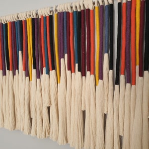 Puede incluir: Un tapiz de macram&eacute; de pared colorido con una variedad de hilos de lana de diferentes colores, incluyendo rojo, naranja, amarillo, verde, azul, morado y negro. Los hilos est&aacute;n atados juntos en la parte superior y cuelgan en cascada de flecos.