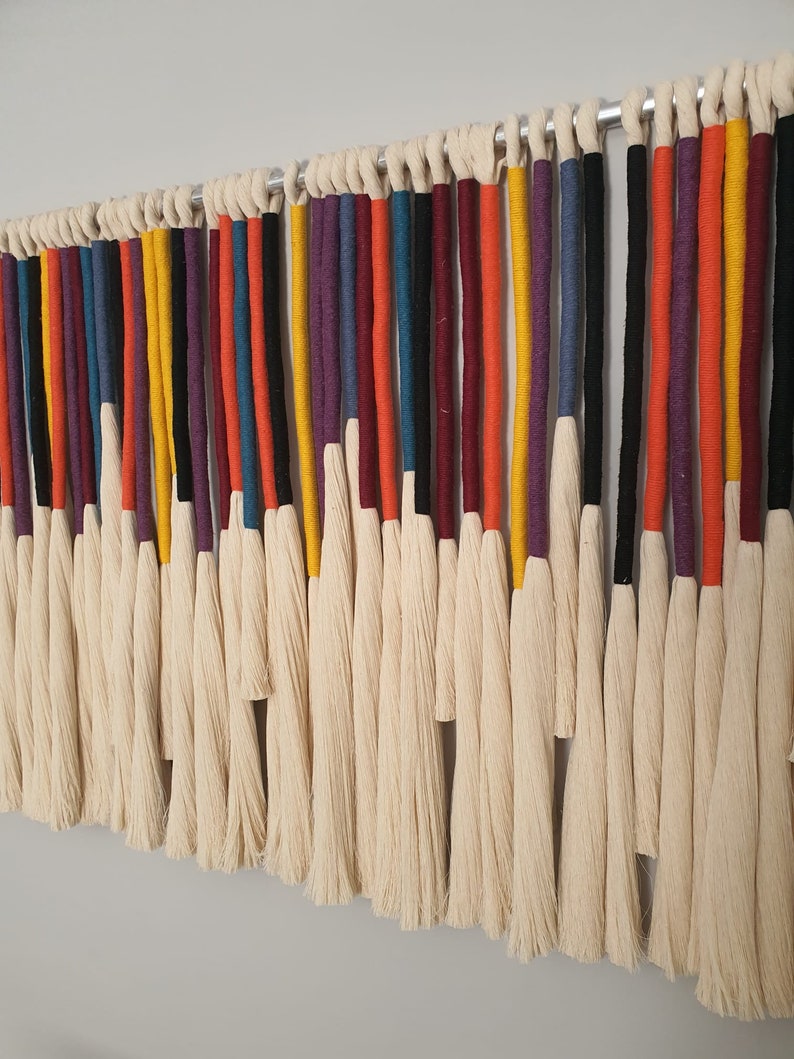 Puede incluir: Un tapiz de macram&eacute; de pared colorido con una variedad de hilos de lana de diferentes colores, incluyendo rojo, naranja, amarillo, verde, azul, morado y negro. Los hilos est&aacute;n atados juntos en la parte superior y cuelgan en cascada de flecos.