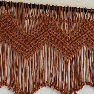May include: A brown macrame wall hanging with a geometric pattern and fringe. The hanging is made with thick cord and has a rustic, bohemian style.