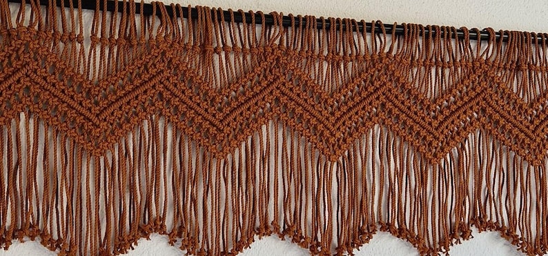 May include: A brown macrame wall hanging with a geometric pattern and fringe. The hanging is made with thick cord and has a rustic, bohemian style.