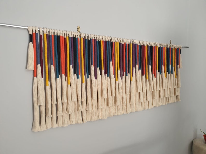 Puede incluir: Un tapiz de macram&eacute; de pared colorido con una variedad de hilos de lana de diferentes colores y borlas blancas. El tapiz est&aacute; hecho con una variedad de colores, incluyendo rojo, naranja, amarillo, verde, azul, morado y negro.