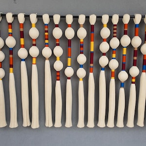 May include: A decorative wall hanging featuring multiple strands of off-white tassels. Each strand has a series of white spheres and is accented with colorful bands of red, orange, yellow, and blue. The piece is suspended from a black rod.
