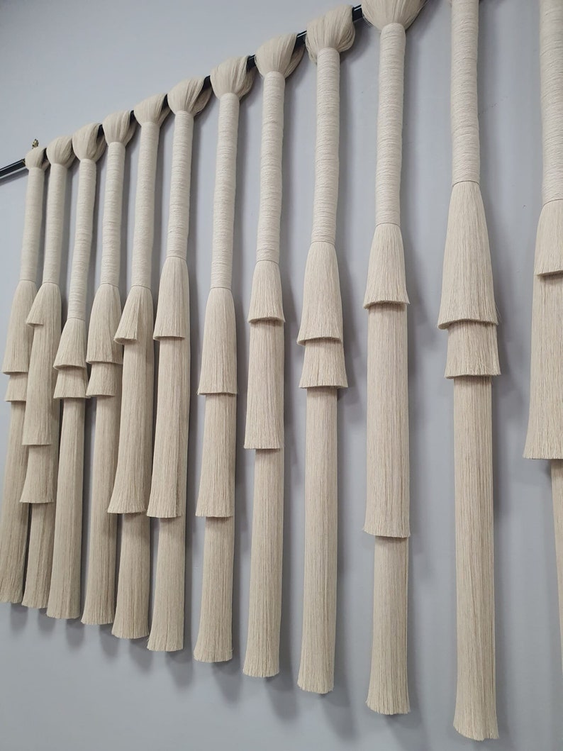 K&ouml;nnte beinhalten: Ein Wandbehang aus Naturfaserseil mit einem mehrstufigen Fransenmuster. Das Seil ist hellbeige und die Fransen sind in einem kaskadierenden Muster angeordnet.