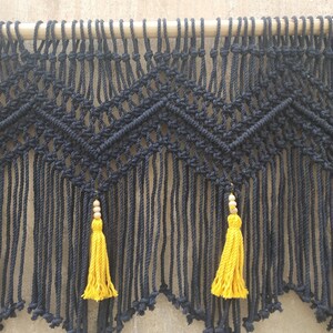 May include: Black macrame wall hanging with a geometric pattern and yellow tassels. The hanging is attached to a wooden dowel.