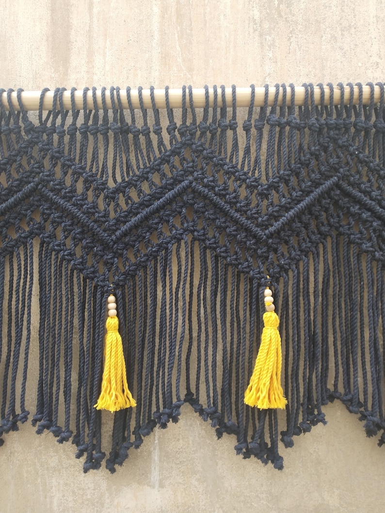 May include: Black macrame wall hanging with a geometric pattern and yellow tassels. The hanging is attached to a wooden dowel.