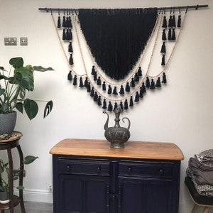 May include: A black macrame wall hanging with multiple strands of black tassels and wooden beads. The hanging is displayed on a white wall above a dark blue dresser with a wooden top.