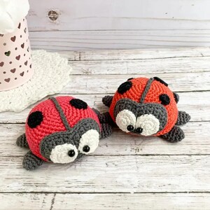 May include: Two crocheted ladybug toys with black spots, red bodies, and gray faces. The ladybugs have white eyes and are sitting on a wooden surface.