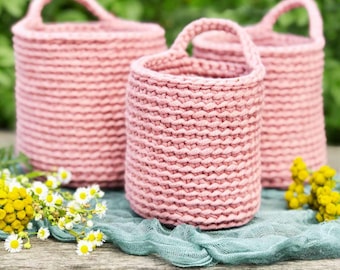 May include: Three pink crocheted baskets with handles. The baskets are made of a thick, textured yarn and have a slightly distressed look. The baskets are sitting on a wooden surface with a light blue fabric underneath.