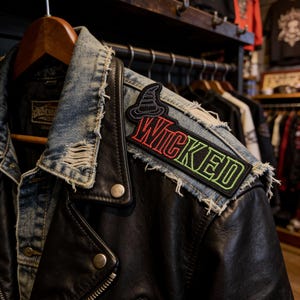 May include: Black leather jacket with a denim collar, displaying a "Wicked" patch with a witch hat design. The patch has red and green lettering. The jacket is on a wooden hanger in a clothing store.