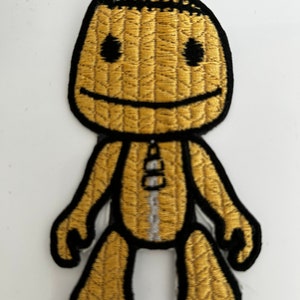 May include: A brown and black embroidered patch of a smiling character with a zipper down the front. The character has a stitched texture and black eyes.