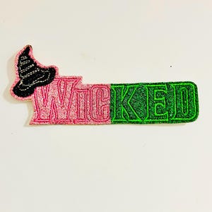 May include: A pink and green glitter iron-on patch with the word "WICKED" and a black witch's hat.