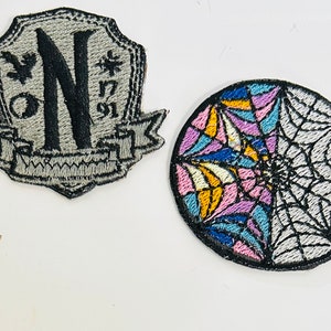 May include: Two embroidered iron-on patches. One is a grey shield with a black letter N and a banner. The other is a round patch with a colourful spiderweb design.