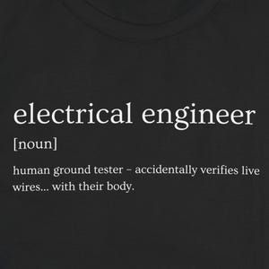 Op de afbeelding: Zwart T-shirt met de definitie van "elektrotechnisch ingenieur" in witte tekst. De definitie bevat het woord "zelfstandig naamwoord" en een humoristische beschrijving van het beroep.