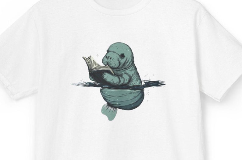 Op de afbeelding: Een wit T-shirt met korte mouwen en een centrale grafische illustratie van een blauwgroene zeekoe die gedeeltelijk in het water is ondergedompeld terwijl deze een open boek vasthoudt en leest. De zeekoe heeft een getextureerde huid.