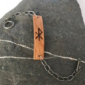 May include: A wooden bracelet with a silver chain and a burned-in symbol. The bracelet is on a gray rock.