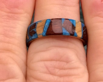 May include: A wooden ring with a blue, brown, and tan inlay pattern. The ring is worn on a finger.
