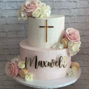 May include: Two-tiered white and pink cake decorated with white and pink flowers, a gold cross, and the name "Maxwell".