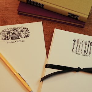 May include: Two sets of personalized stationery with black ink. One set features a floral design with a basket and the name "Evelyn Clifford". The other set features a row of silverware with the same name.