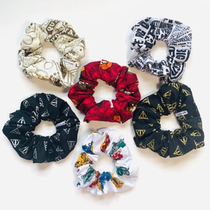 May include: Six different hair scrunchies with various Harry Potter themed prints. The scrunchies are made of fabric and have a gathered elastic band. The prints include a black and white print with the Hogwarts crest, a white print with a colourful illustration of Harry Potter characters, a black print with the Deathly Hallows symbol, a white print with the words "Always" and a heart, a brown print with the words "Mischief Managed" and a black and white print with the words "I solemnly swear that I am up to no good."