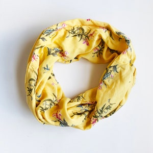May include: A yellow infinity scarf with a floral pattern. The scarf is a bright yellow color with a repeating pattern of green leaves and pink and blue flowers. The scarf is made of a soft, lightweight fabric.